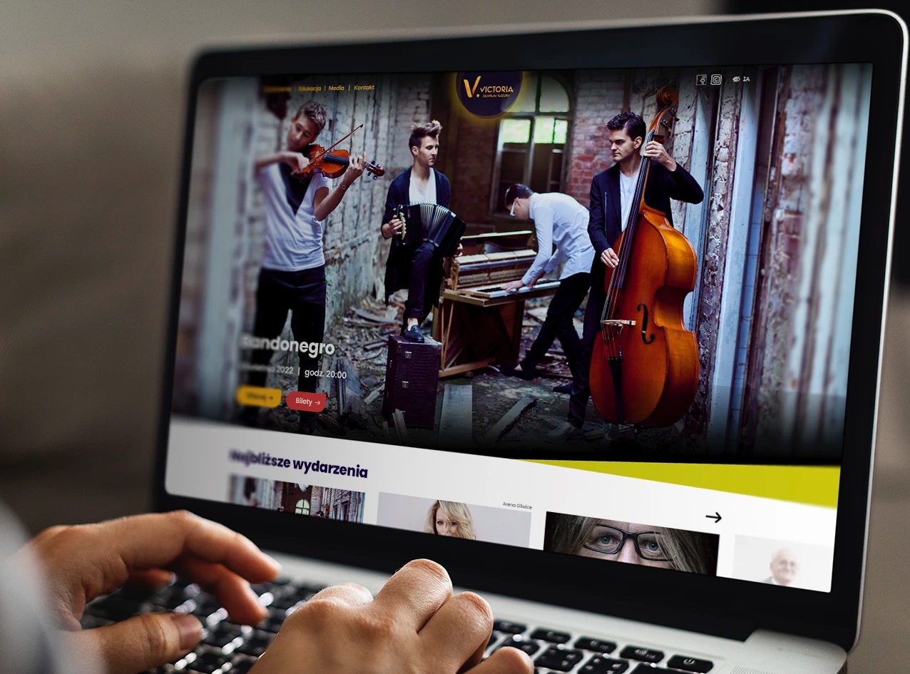 Ekran laptopa wyświetlający stronę internetową zespołu muzycznego z czterema muzykami grającymi na skrzypcach, akordeonie, pianinie i kontrabasie na tle starej, ceglanej ściany; dłonie użytkownika...