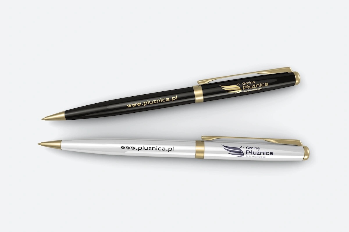Dwa długopisy z nadrukiem logo i adresu strony internetowej Gminy Płużnica, czarny i biały, leżące na jasnym tle.
