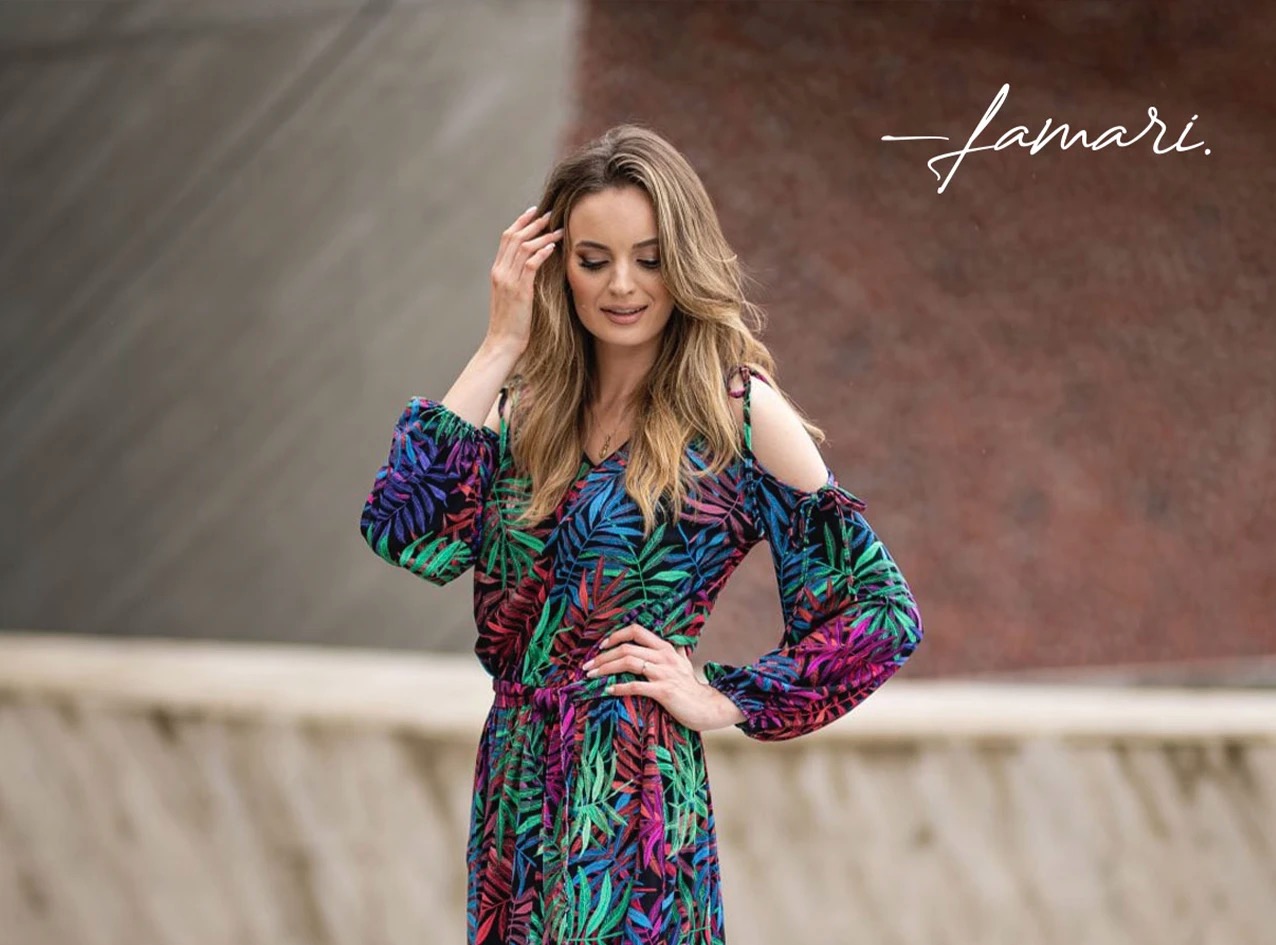 Kobieta w sukience z motywem tropikalnych liści, pozująca na tle betonowej ściany, z widocznym logo marki odzieżowej w prawym górnym rogu.