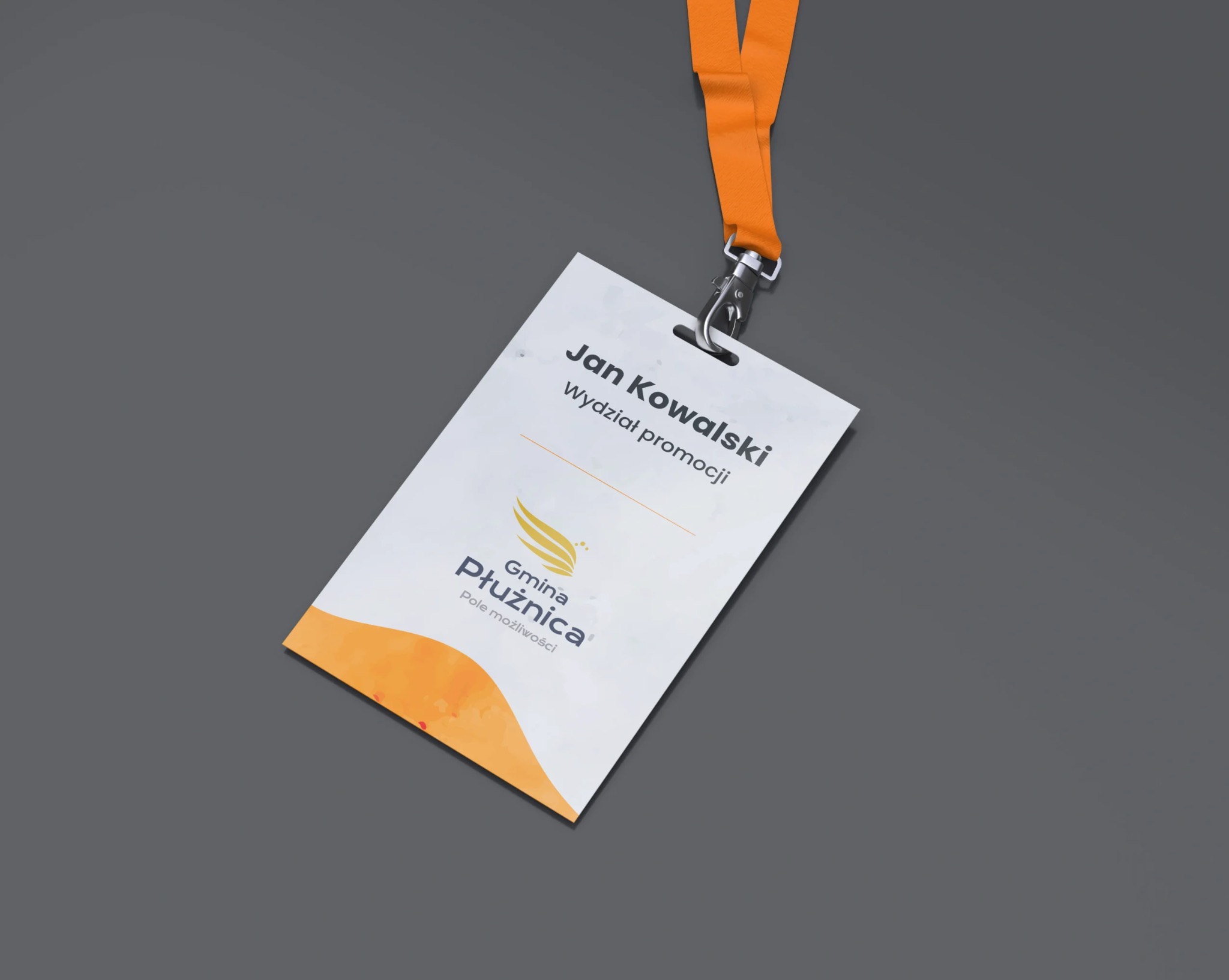 Identyfikator wizualny z logo i danymi osobowymi, zawieszony na pomarańczowej smyczy, leżący na szarym tle.