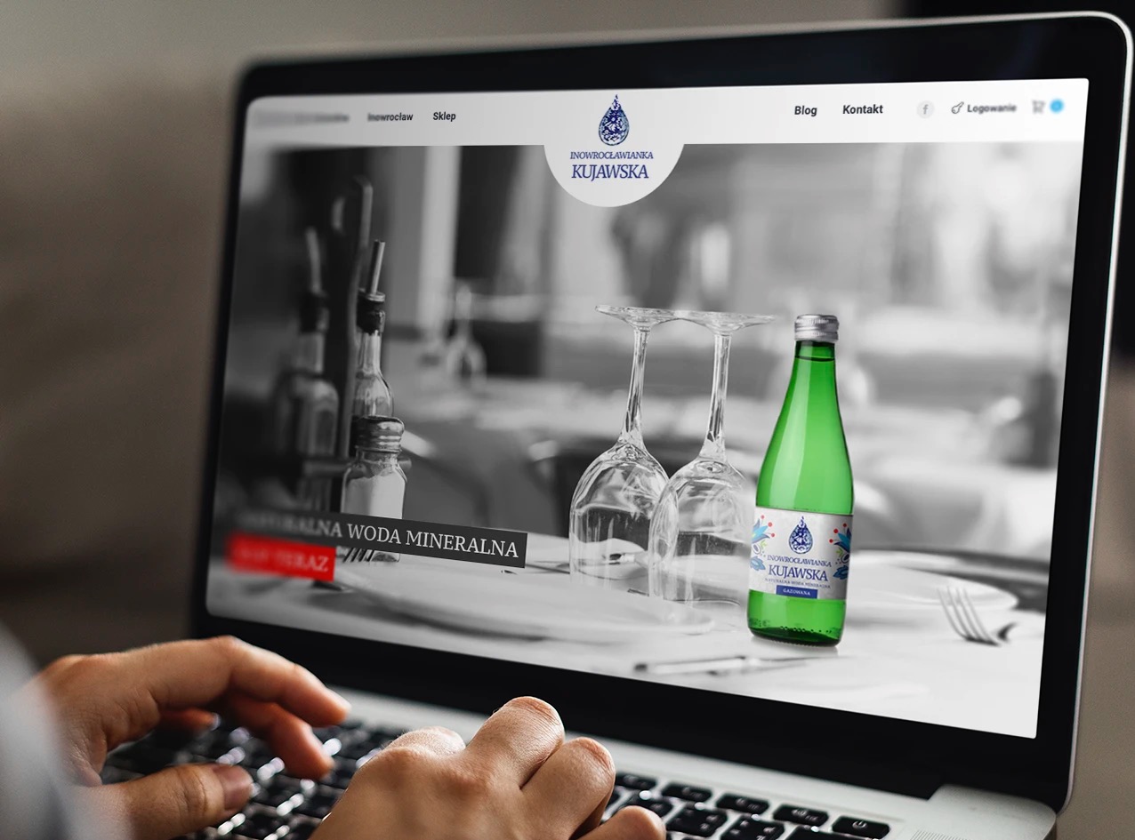 Osoba pisze na laptopie, na ekranie strona internetowa z wizerunkiem butelki wody mineralnej i kieliszków na stole w restauracji.