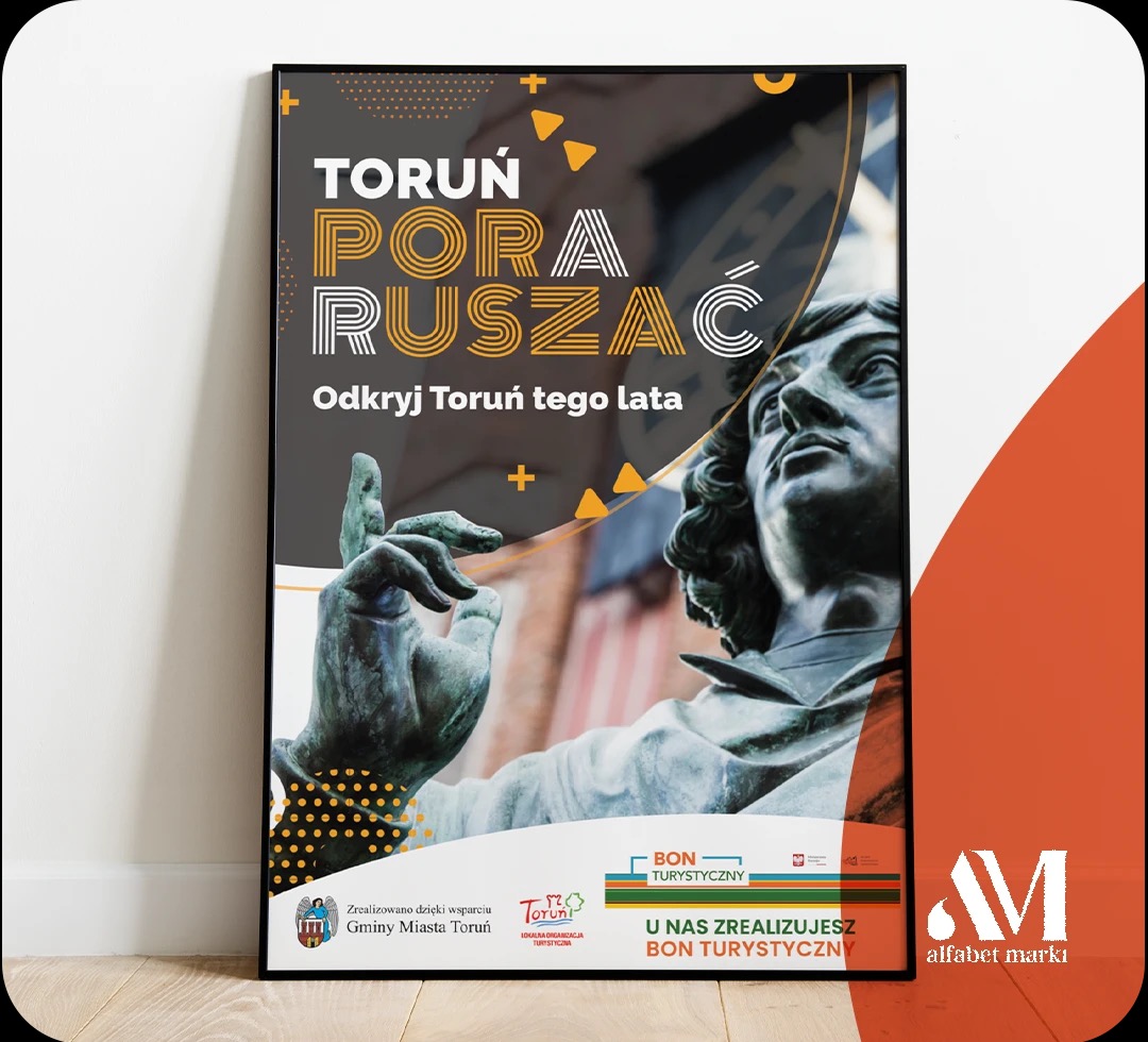 Plakat reklamowy z hasłem 'Toruń Pora Ruszać' i zdjęciem pomnika Mikołaja Kopernika, zachęcający do odkrywania Torunia tego lata, w czarnej ramie.