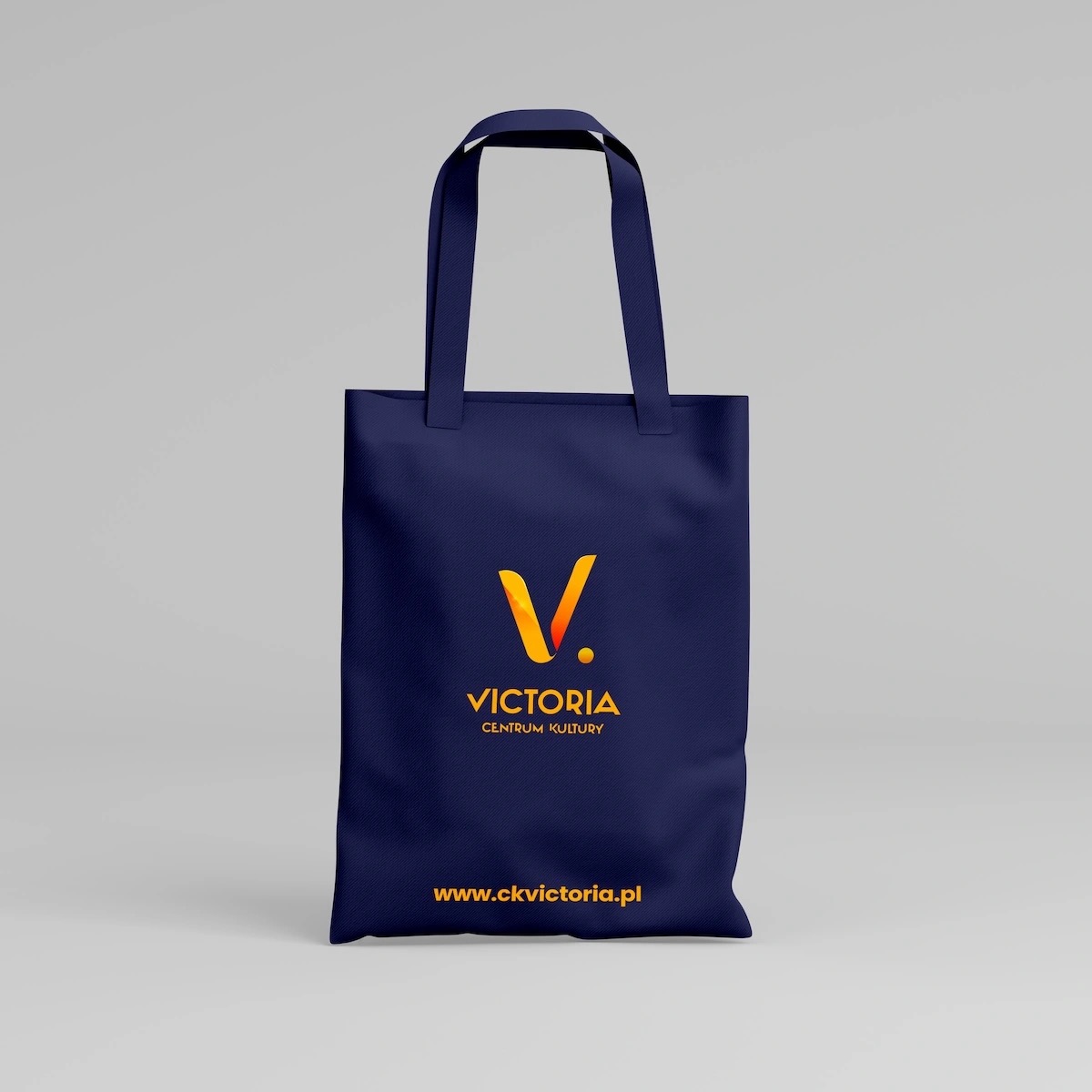 Granatowa torba bawełniana z pomarańczowym logo Victoria Centrum Kultury i adresem strony internetowej, na jasnoszarym tle.