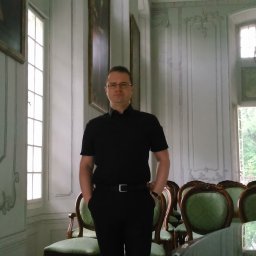 Kancelaria Radcy Prawnego Marcin Gruszecki - Mężczyzna w czarnym stroju stoi w eleganckim wnętrzu z dekoracyjnymi ścianami, żyrandolem i krzesłami z zielonym obiciem. Portret w reprezentacyjnym otoczeniu.