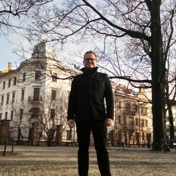 Kancelaria Radcy Prawnego Marcin Gruszecki - Mężczyzna w czarnym płaszczu i okularach stoi na tle klasycystycznej architektury i drzew bez liści w słoneczny dzień. Ujęcie od dołu.