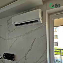 Duoeco Sp. z o.o. - Biały klimatyzator marki Kaisai zamontowany na ścianie z marmurowym wzorem, widoczny uchwyt do telewizora i okno z roletą.