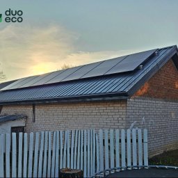 Duoeco Sp. z o.o. - Instalacja paneli fotowoltaicznych na blaszanym dachu budynku z białej cegły w Krakowie, widoczne logo firmy Duo Eco, biały płot i fragment trampoliny w tle.