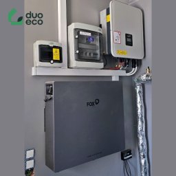 Duoeco Sp. z o.o. - Szara bateria magazynu energii Fox ESS zamontowana na ścianie obok zabezpieczonej rury, z rozdzielnicą elektryczną i falownikiem fotowoltaicznym powyżej, widoczne kable i elementy instalacji...