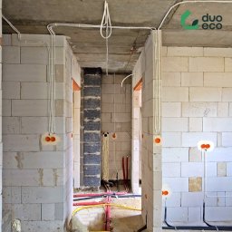 Duoeco Sp. z o.o. - Surowy stan: instalacja elektryczna i rury w ścianach z betonu komórkowego. Widoczne kable w peszlach i puszki instalacyjne. Logo firmy w prawym górnym rogu.