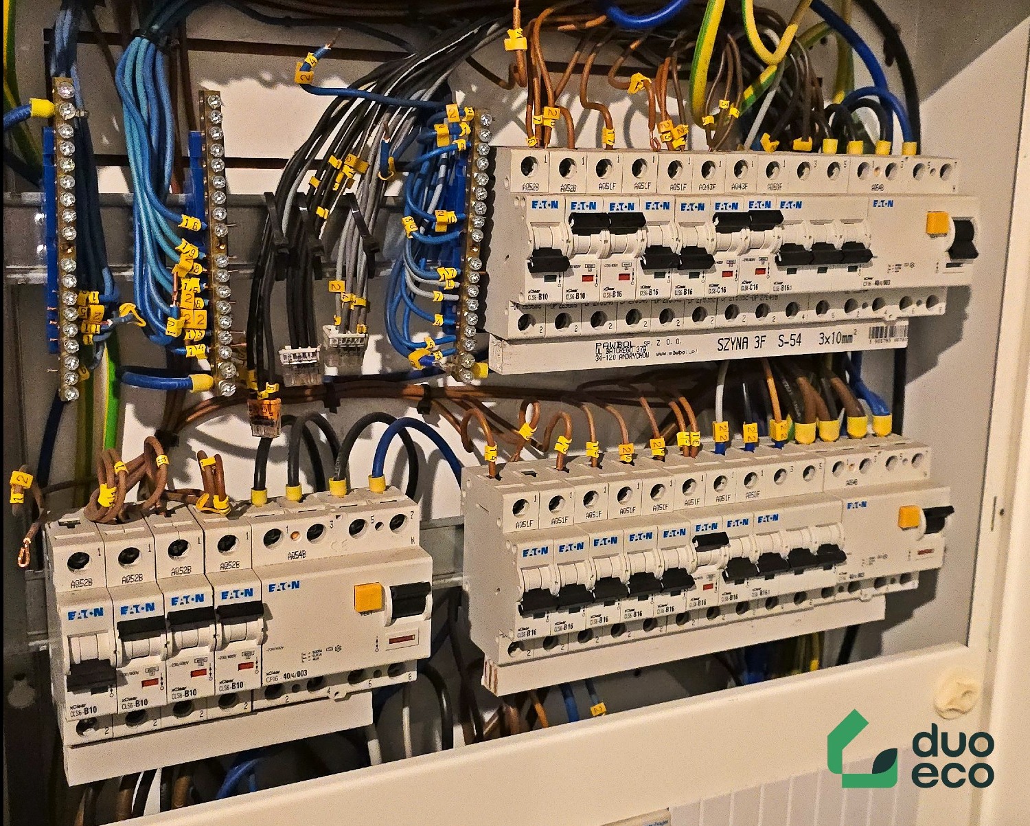 Rozdzielnica elektryczna z bezpiecznikami EATON, oznaczonymi przewodami i żółtymi numerowanymi oznaczeniami, widok na zamontowaną instalację elektryczną wewnątrz obudowy.