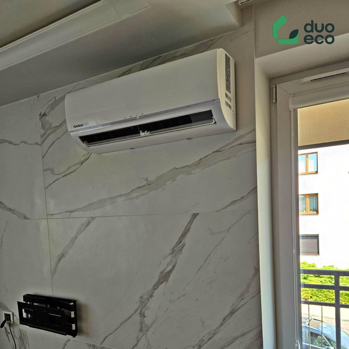 Biały klimatyzator marki Kaisai zamontowany na ścianie z marmurowym wzorem, widoczny uchwyt do telewizora i okno z roletą.