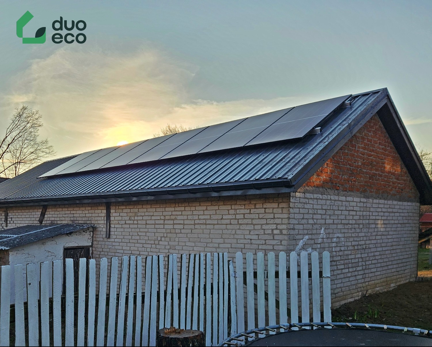 Instalacja paneli fotowoltaicznych na blaszanym dachu budynku z białej cegły w Krakowie, widoczne logo firmy Duo Eco, biały płot i fragment trampoliny w tle.