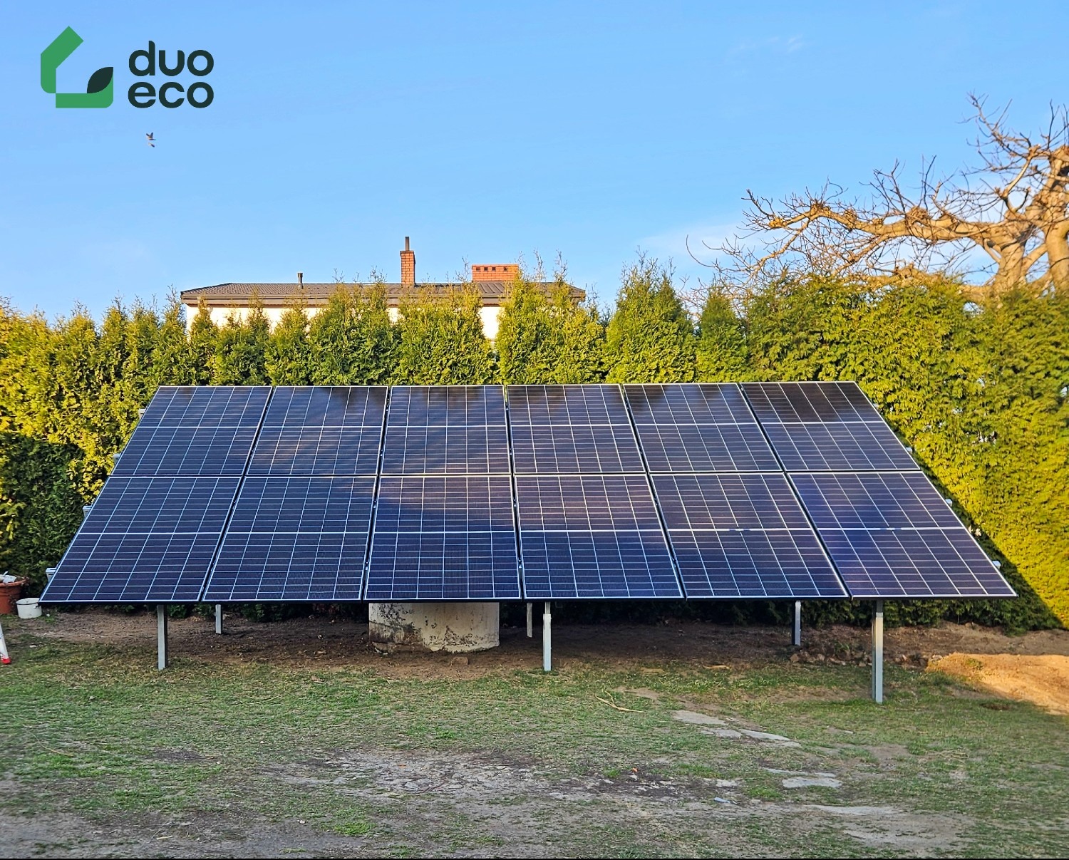 Panel fotowoltaiczny na konstrukcji wsporczej z widocznym logo firmy duo eco w tle zielone tuje i dom jednorodzinny.