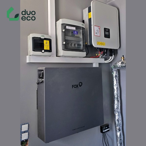 Szara bateria magazynu energii Fox ESS zamontowana na ścianie obok zabezpieczonej rury, z rozdzielnicą elektryczną i falownikiem fotowoltaicznym powyżej, widoczne kable i elementy instalacji...