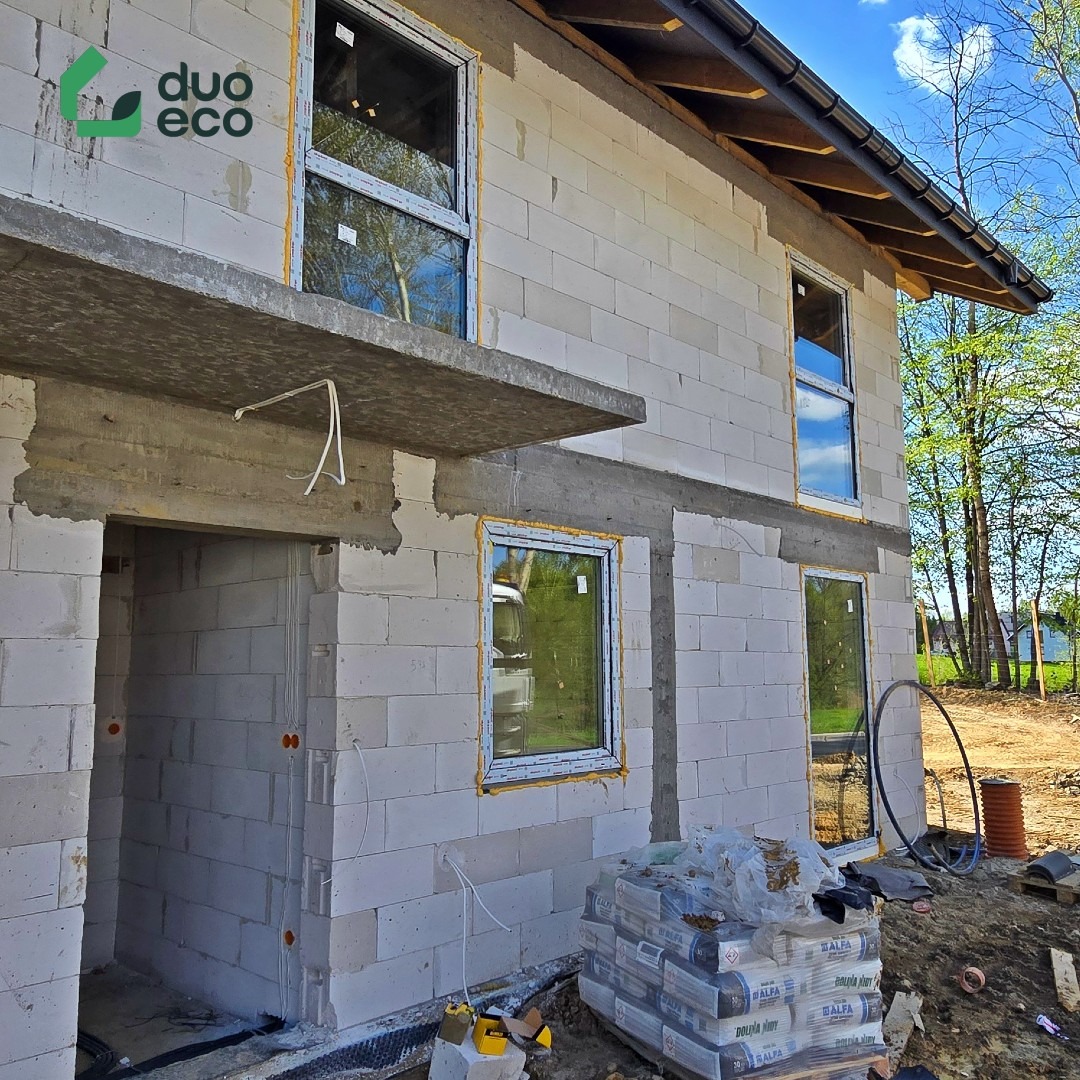 Surowy stan domu z betonu komórkowego z oknami i widocznymi przewodami elektrycznymi. Na pierwszym planie paleta z zaprawą murarską.