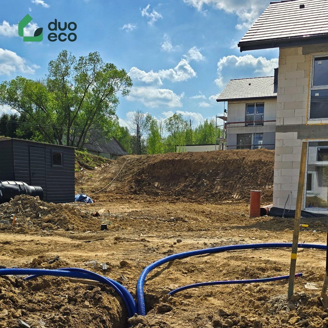 Niebieskie rury w wykopie na placu budowy z domami w tle, w słoneczny dzień. Widoczny fragment instalacji i ziemia.