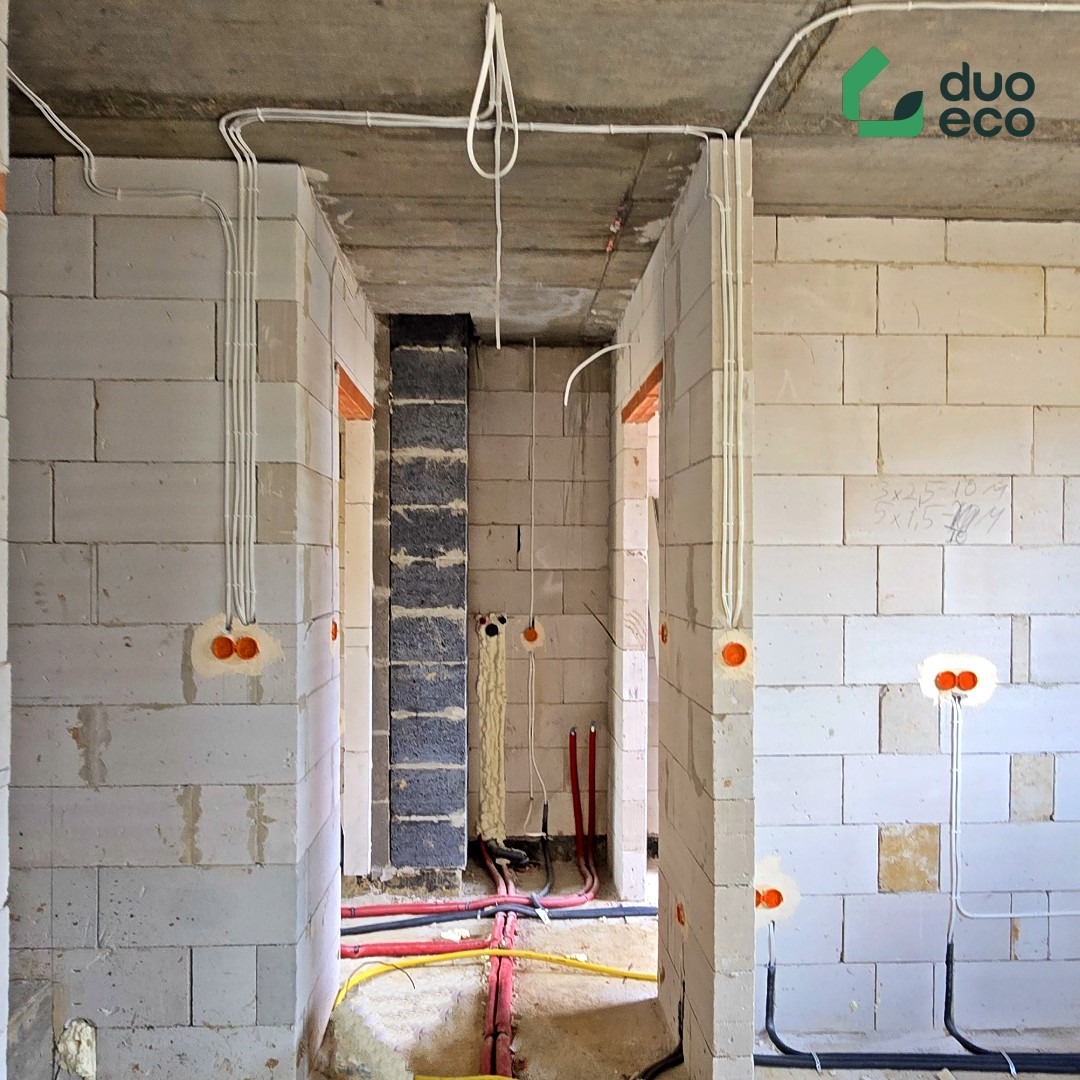 Surowy stan: instalacja elektryczna i rury w ścianach z betonu komórkowego. Widoczne kable w peszlach i puszki instalacyjne. Logo firmy w prawym górnym rogu.