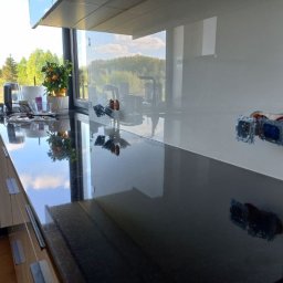 SWEDA HOUSING SPÓŁKA Z OGRANICZONĄ ODPOWIEDZIALNOŚCIĄ - Biała ściana nad blatem kuchennym z zamontowanymi gniazdkami, odbijająca się w błyszczącej powierzchni blatu. Widok na zewnątrz przez okno.