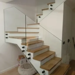 Nowoczesne schody z drewnianymi stopniami i szklaną balustradą, mocowaną na czarnych elementach, w jasnym wnętrzu w Pruszczu Gdańskim.