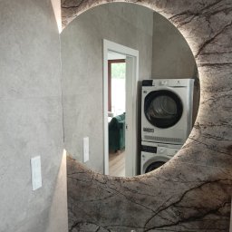 SWEDA HOUSING SPÓŁKA Z OGRANICZONĄ ODPOWIEDZIALNOŚCIĄ - Okrągłe lustro z podświetleniem LED i marmurkowym wzorem, odbijające pralkę i suszarkę w nowoczesnej łazience z płytkami w odcieniach szarości.