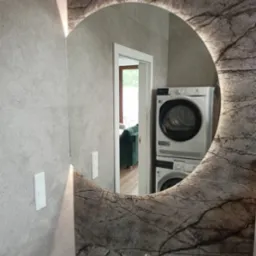 Okrągłe lustro z podświetleniem LED i marmurkowym wzorem, odbijające pralkę i suszarkę w nowoczesnej łazience z płytkami w odcieniach szarości.