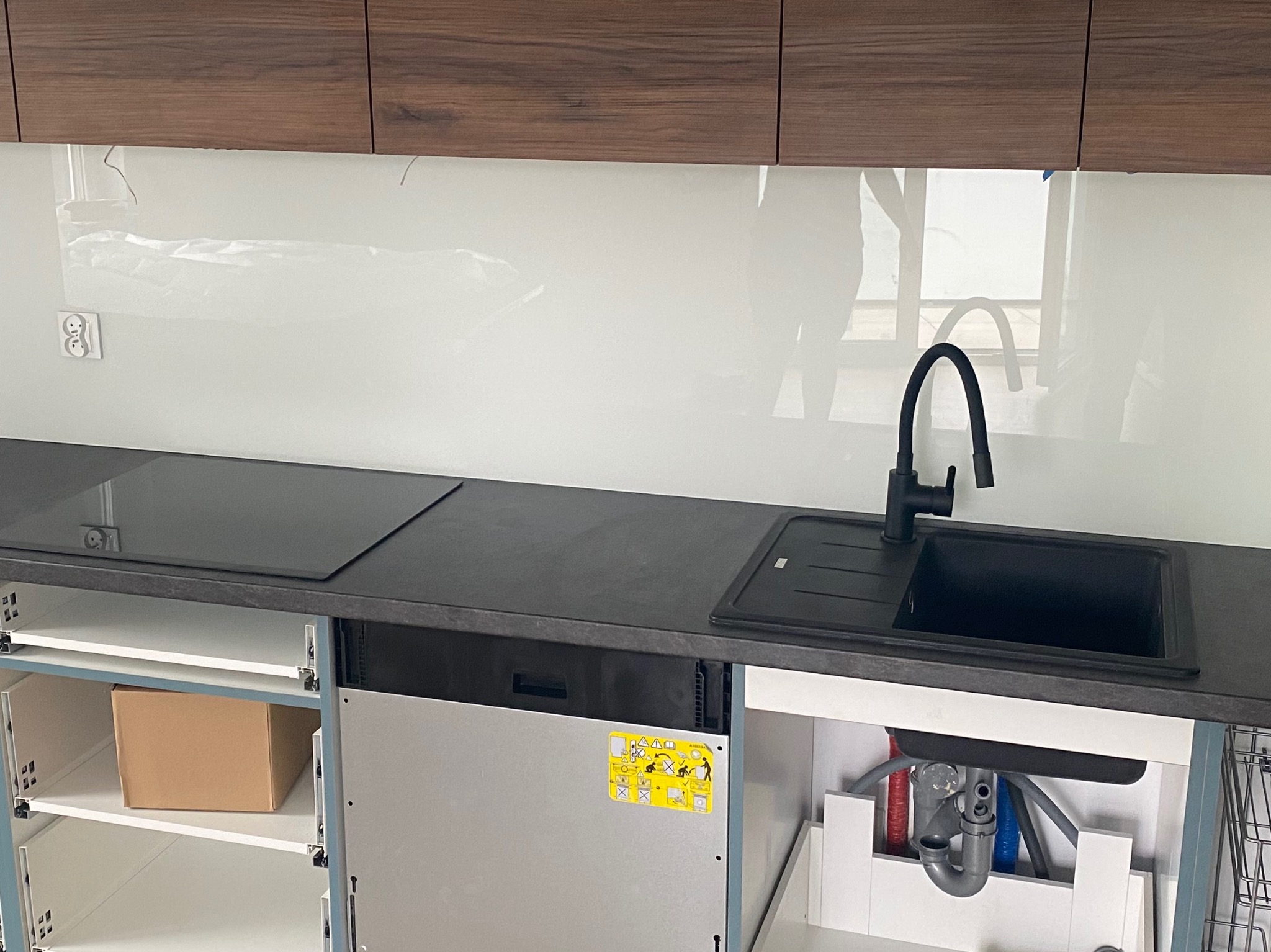 Kuchnia w trakcie montażu: widoczny szklany panel nad blatem, czarny zlew i płyta grzewcza. Szafki bez frontów, ukazują instalacje i karton w środku.