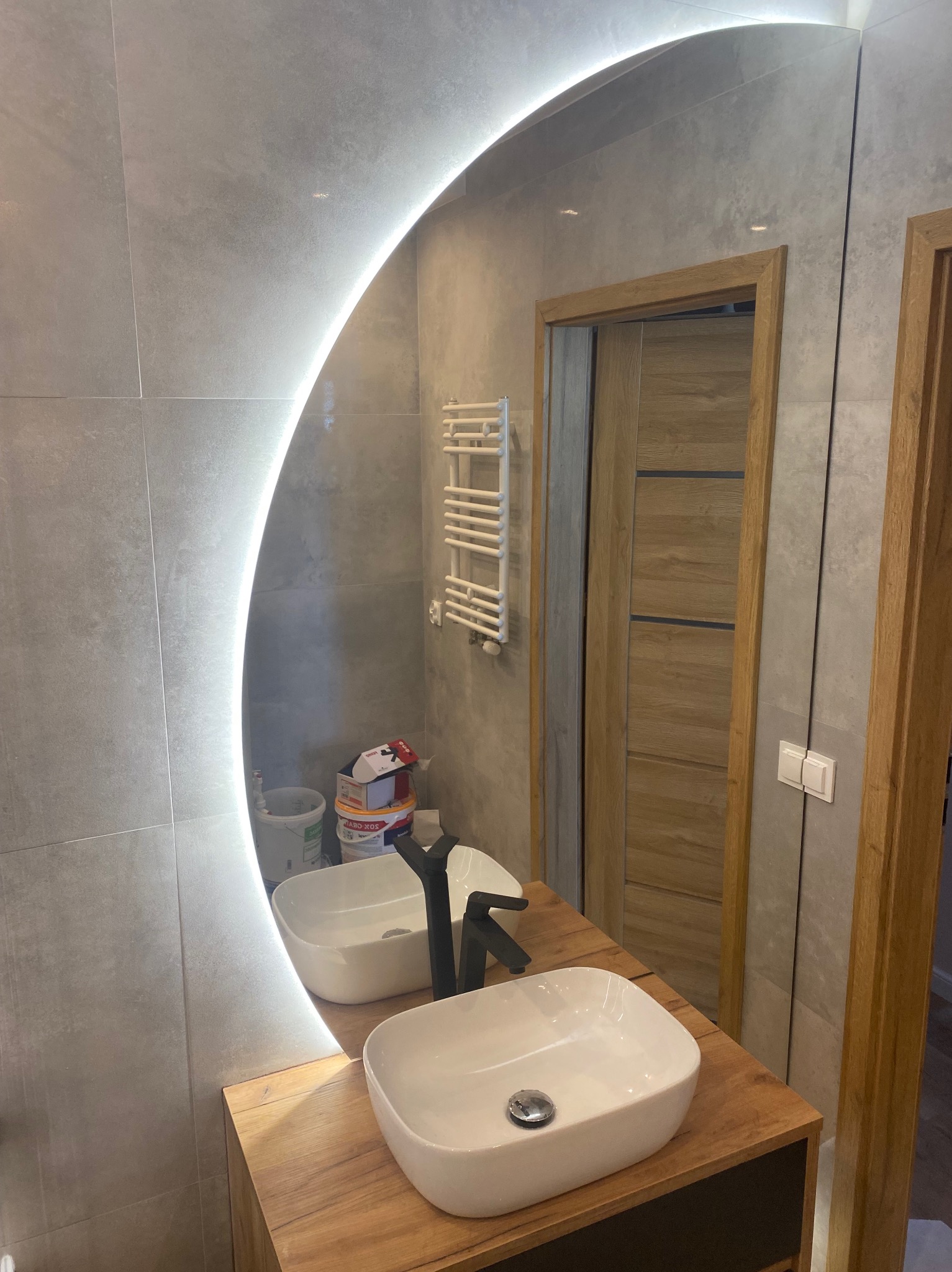 Owalne lustro z podświetleniem LED w nowoczesnej łazience z szarą płytką i drewnianym blatem pod umywalką, odbijające drzwi i grzejnik.