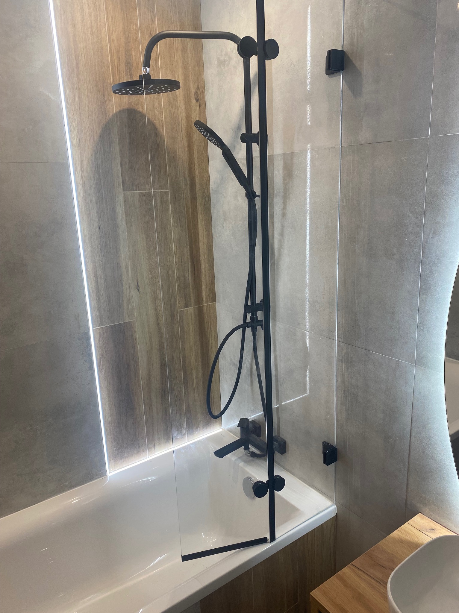 Minimalistyczna kabina prysznicowa ze szklanym panelem, czarną armaturą i subtelnym oświetleniem LED w nowoczesnej łazience z płytkami imitującymi drewno.