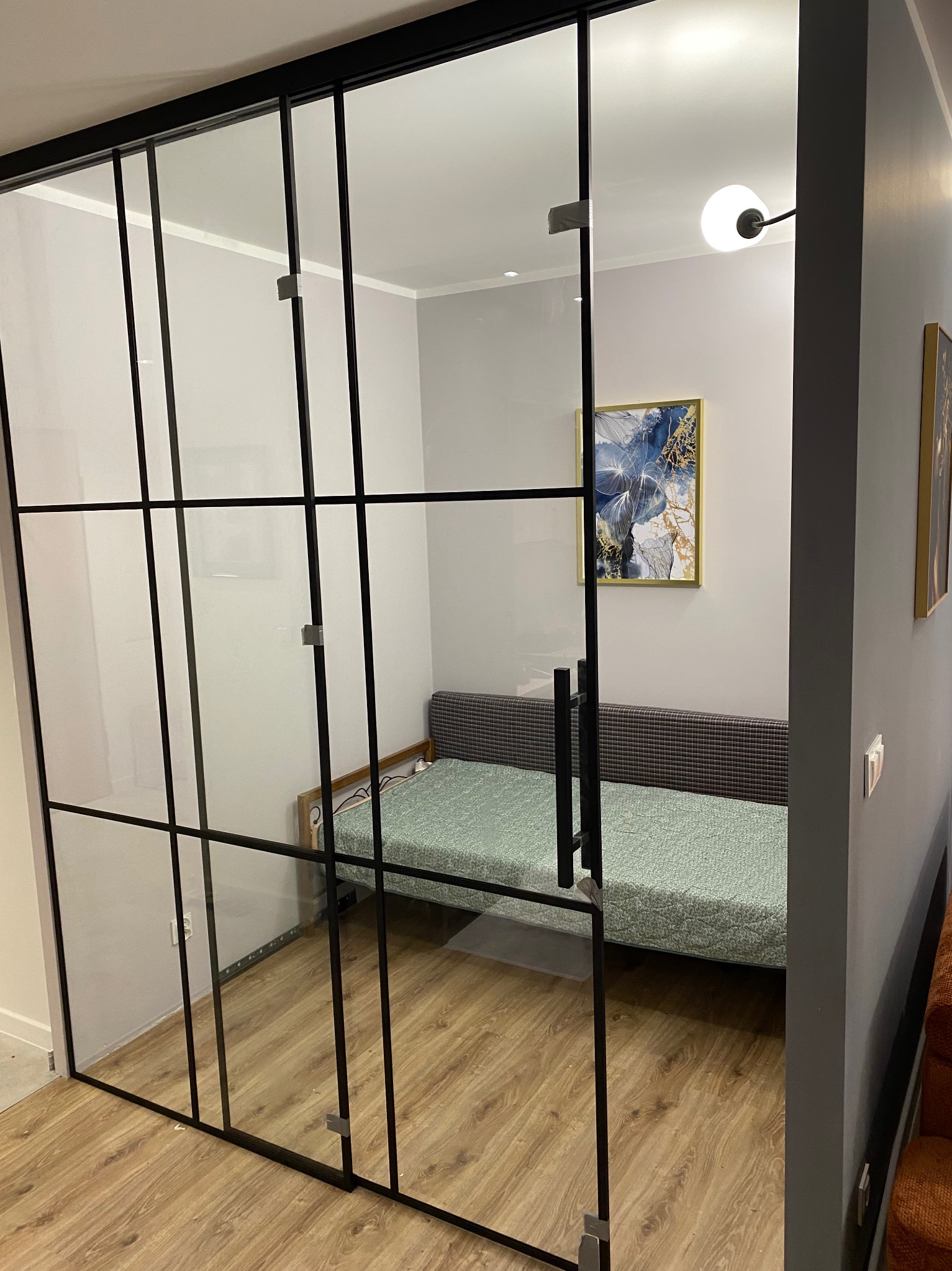 Nowoczesne drzwi szklane z czarnymi szprosami oddzielają pokój z kanapą. Widoczne detale mocowań i rama drzwi. Podłoga drewniana, na ścianie obraz.