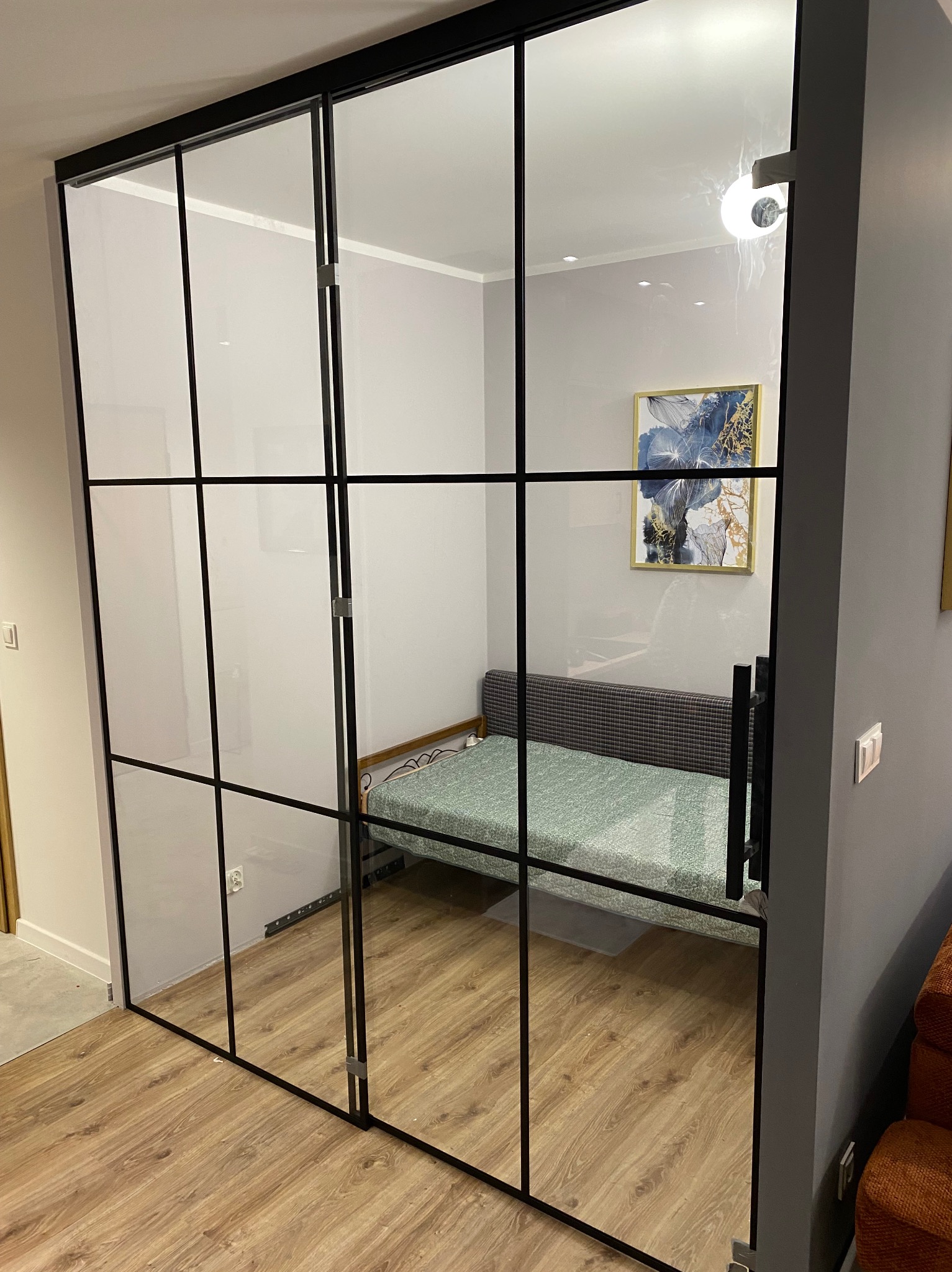 Nowoczesne, przesuwne drzwi szklane z czarnymi szprosami oddzielają pokój. Widoczny fragment wnętrza z łóżkiem i obrazem na ścianie. Minimalistyczny design.