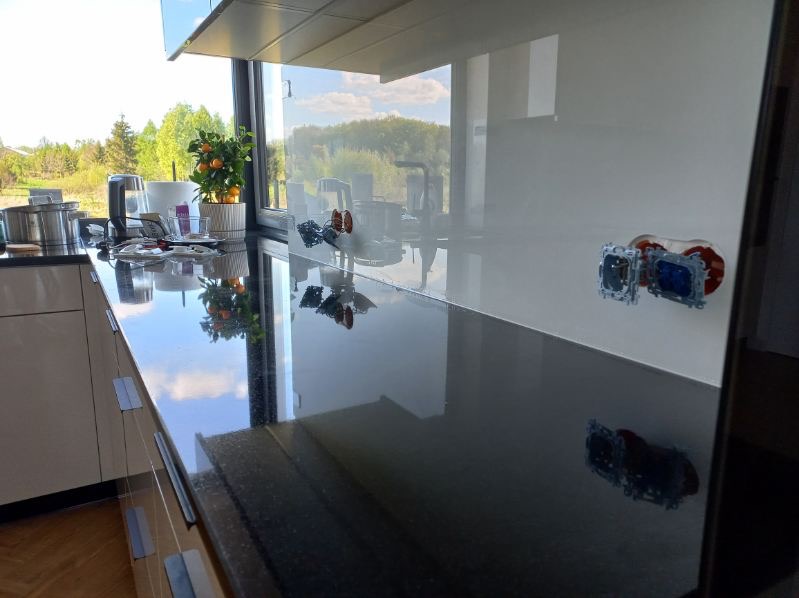 Biała ściana nad blatem kuchennym z zamontowanymi gniazdkami, odbijająca się w błyszczącej powierzchni blatu. Widok na zewnątrz przez okno.