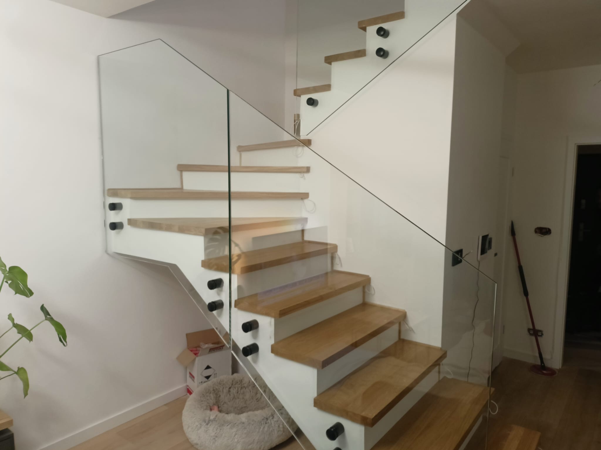 Nowoczesne schody z drewnianymi stopniami i szklaną balustradą, mocowaną na czarnych elementach, w jasnym wnętrzu w Pruszczu Gdańskim.