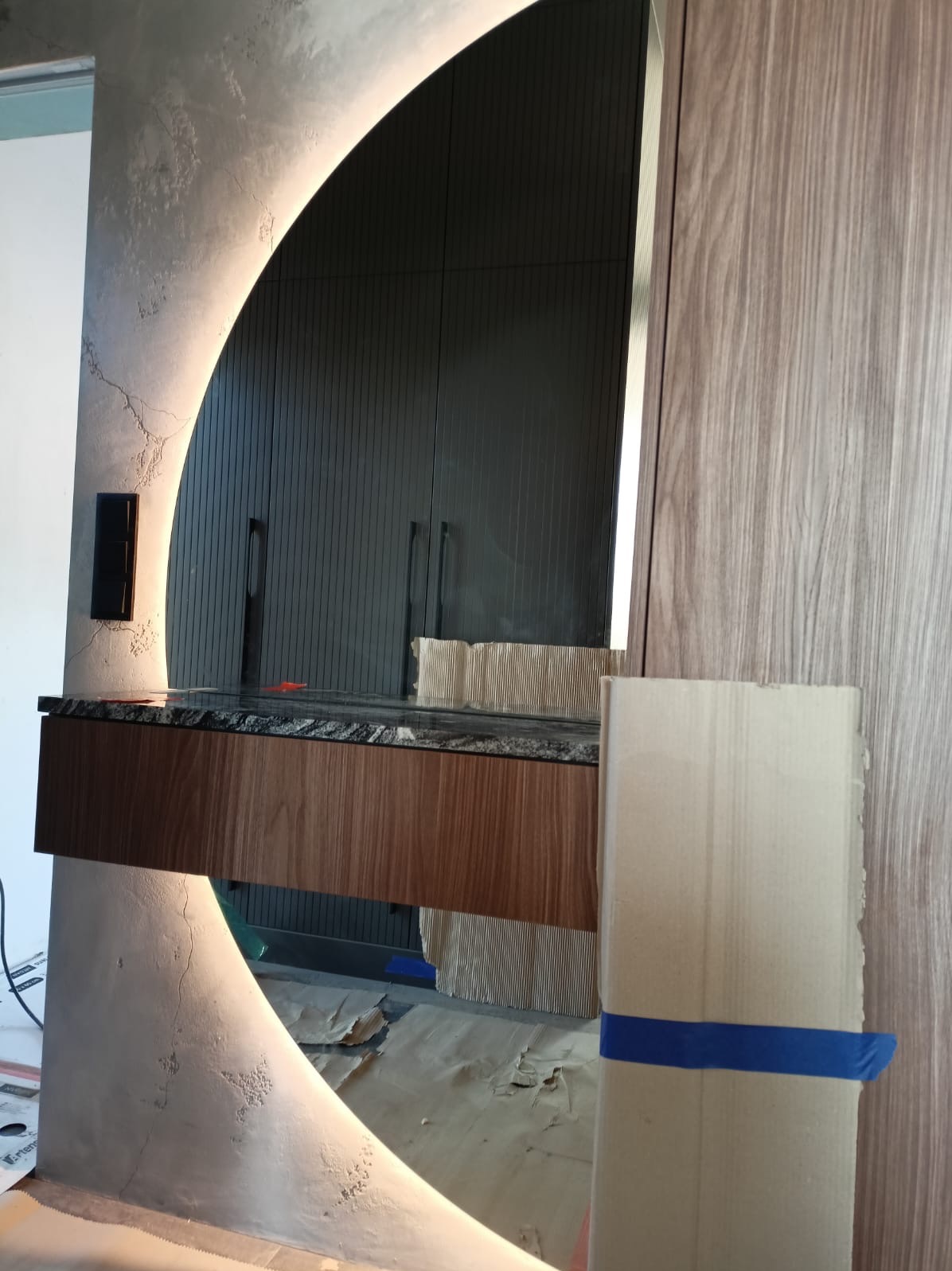 Nowoczesna toaletka z podświetlanym lustrem w kształcie łuku, marmurowym blatem i drewnianą szafką, w trakcie montażu, zabezpieczona kartonem.