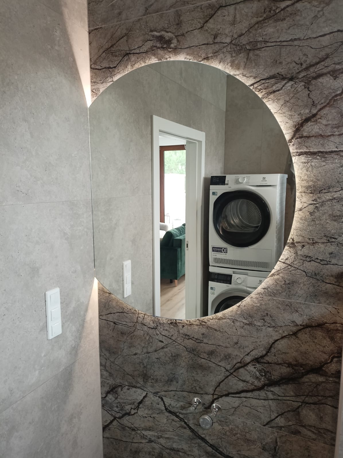 Okrągłe lustro z podświetleniem LED i marmurkowym wzorem, odbijające pralkę i suszarkę w nowoczesnej łazience z płytkami w odcieniach szarości.