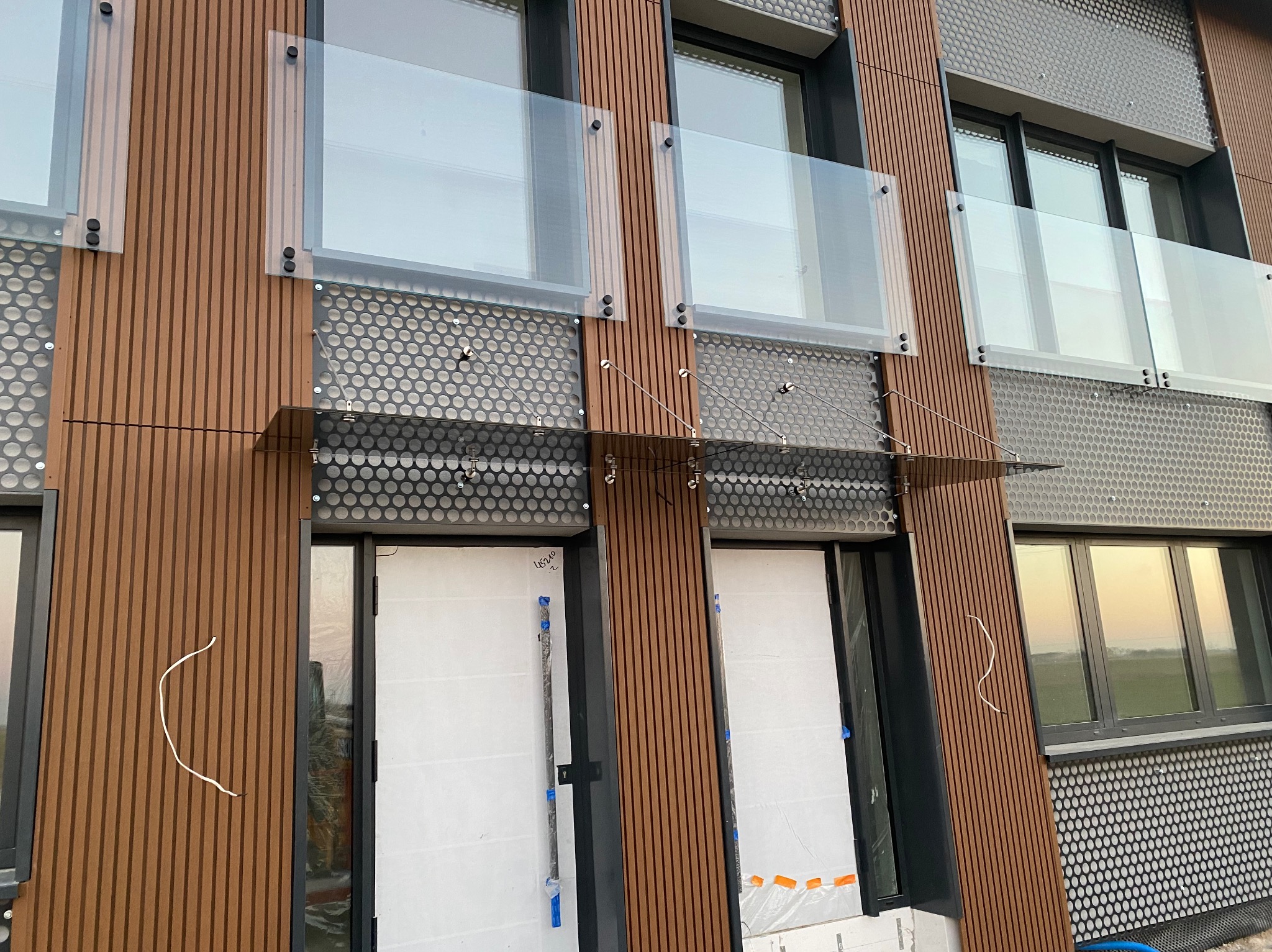 Elewacja budynku z balkonami z przezroczystego szkła, wspartymi na metalowych elementach. Fasada łączy drewniane panele i perforowaną blachę.