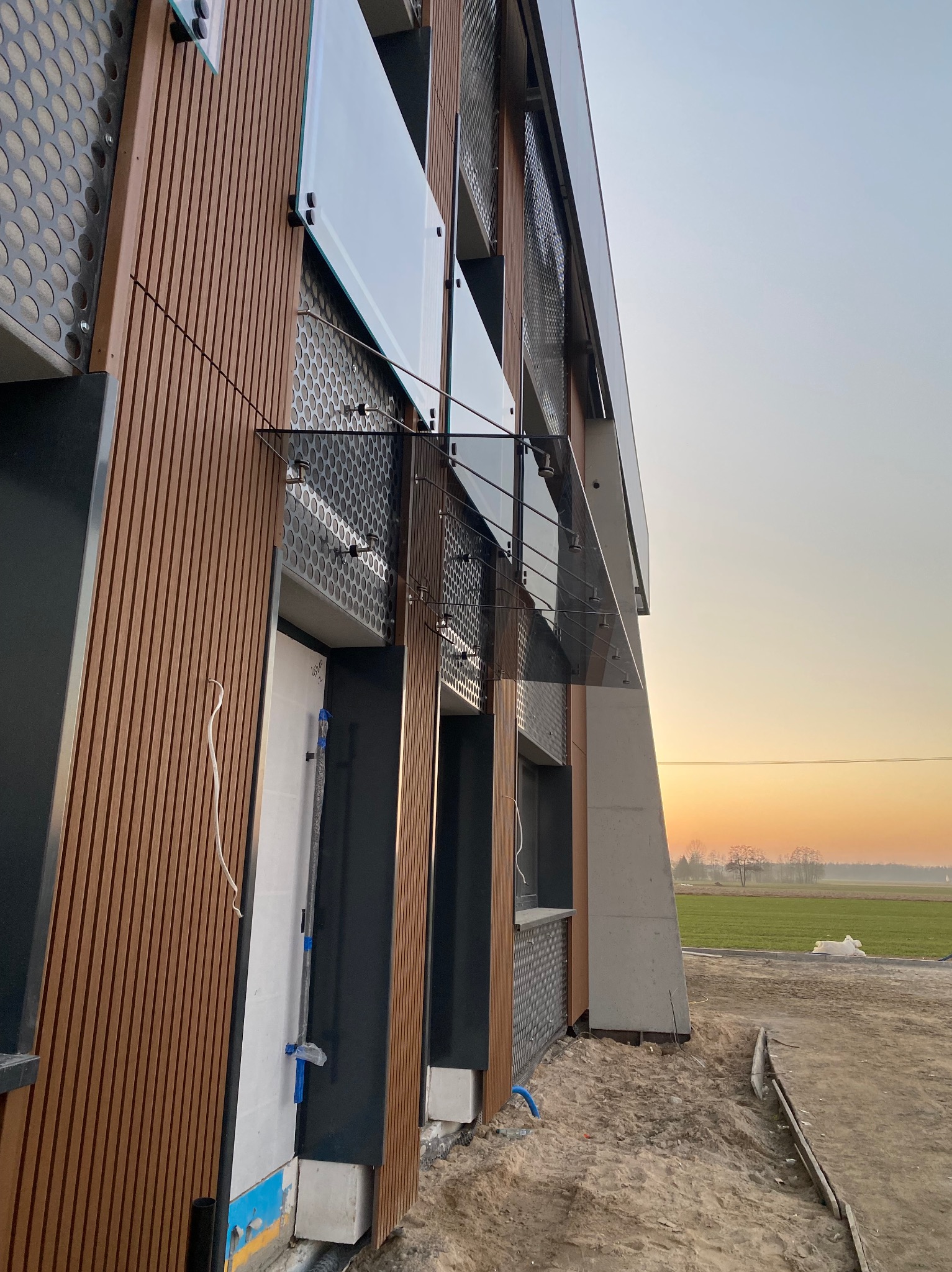 Nowoczesna fasada budynku z drewnianymi panelami i szklanymi elementami, widok z dołu pod kątem, na tle zachodzącego słońca i zielonego pola.