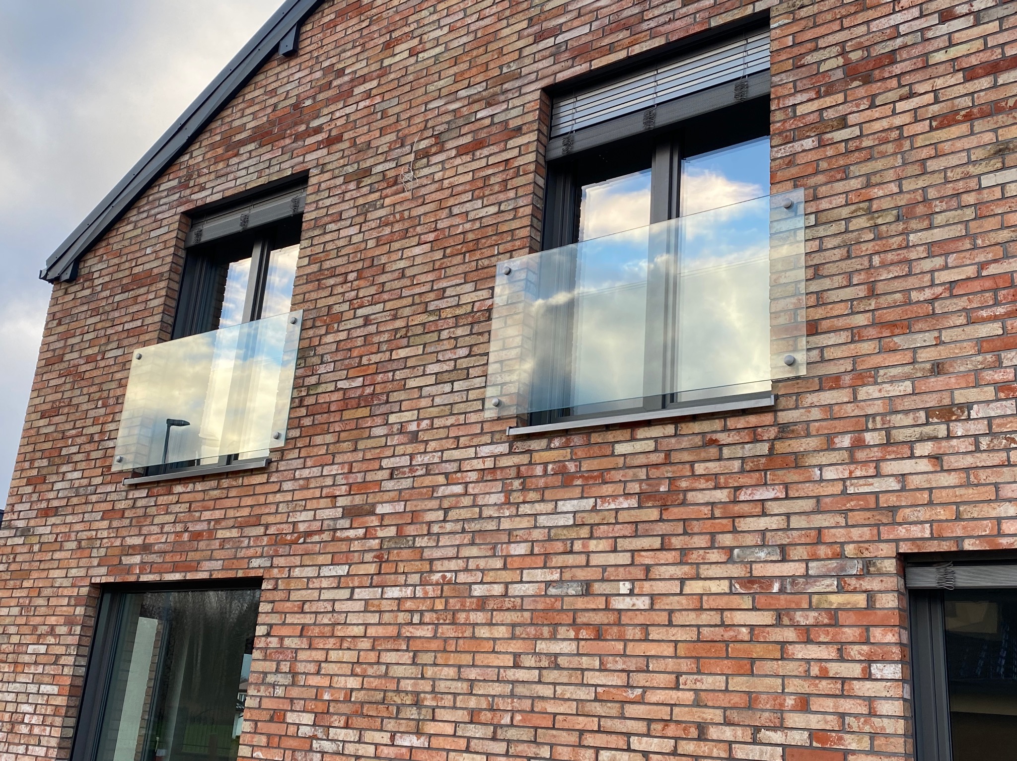 Ceglana fasada domu z oknami i szklanymi balustradami balkonowymi, w których odbija się niebo. Widok z dołu, podkreślający nowoczesny design.