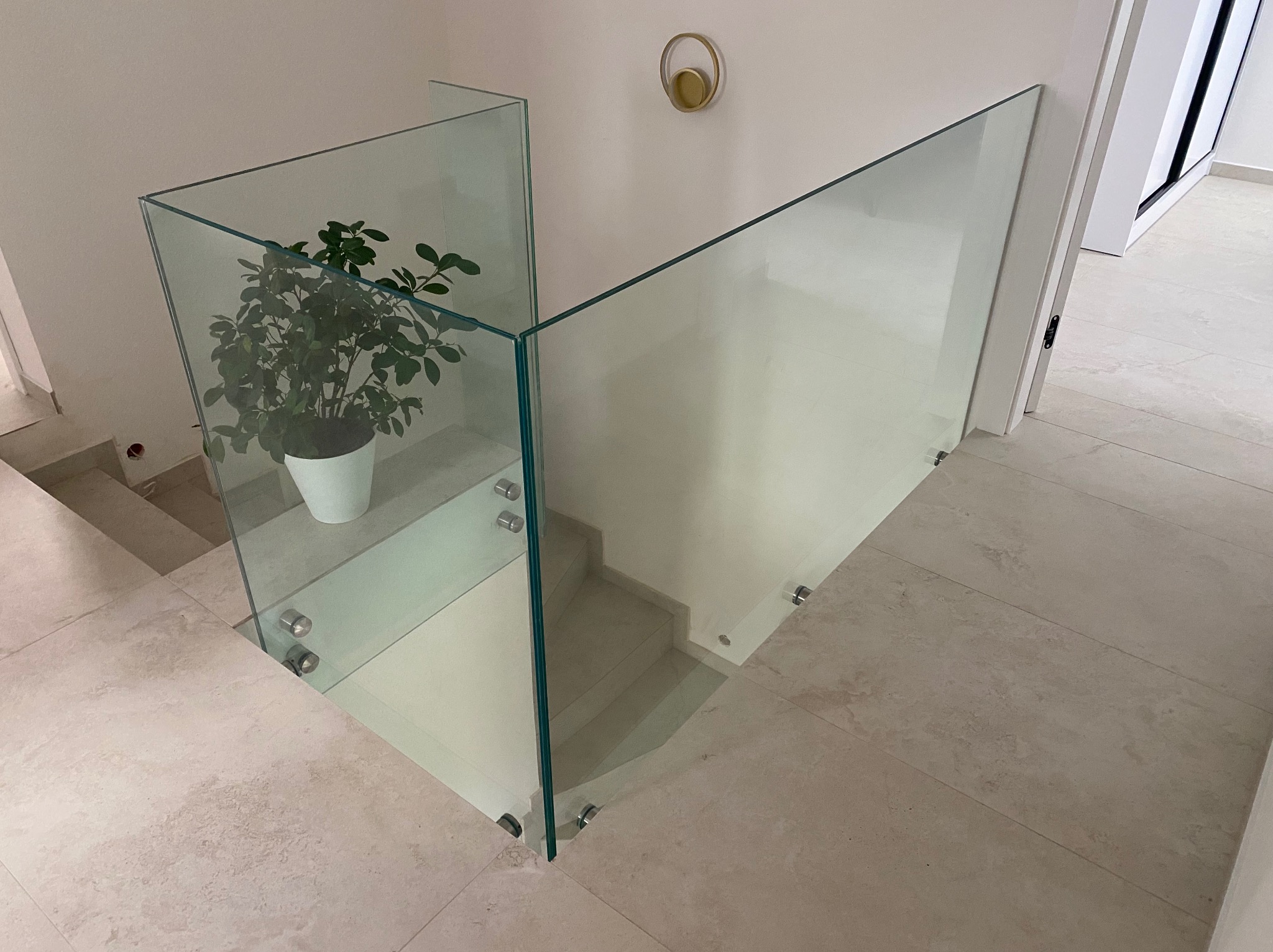 Nowoczesna, minimalistyczna szklana balustrada schodowa z rośliną w doniczce, mocowana punktowo, oświetlona złotym kinkietem. Jasne płytki podłogowe.