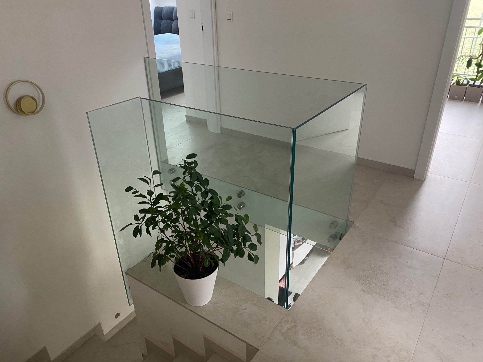 Nowoczesna, minimalistyczna szklana balustrada schodowa z rośliną doniczkową. Jasne wnętrze, neutralna kolorystyka, widok z góry na schody i fragment pokoju.