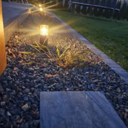 Oświetlony fragment ogrodu z kamieniami ozdobnymi, trawą ozdobną i pojedynczą płytą chodnikową, w tle trampolina i drzewa.