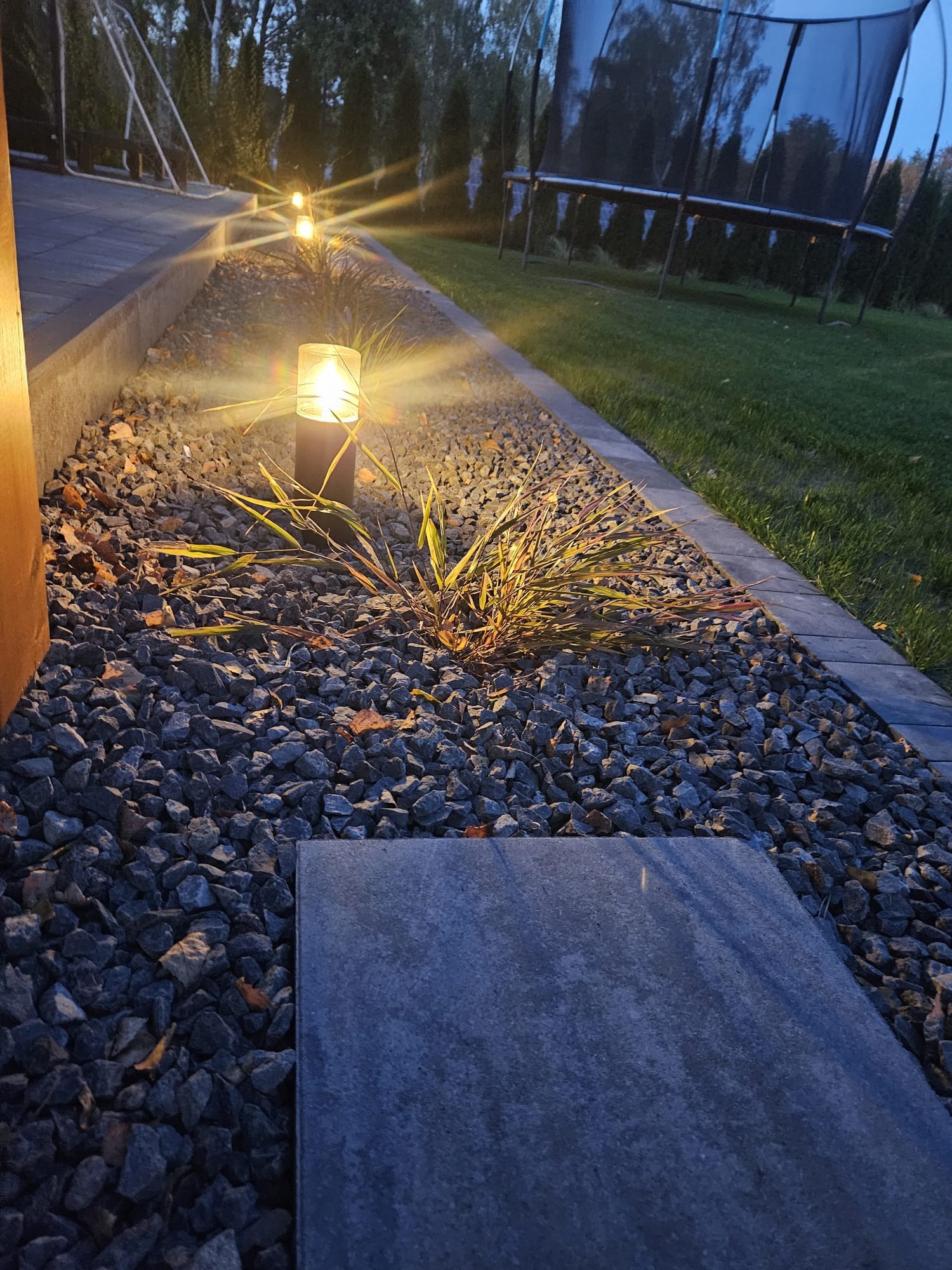 Oświetlony fragment ogrodu z kamieniami ozdobnymi, trawą ozdobną i pojedynczą płytą chodnikową, w tle trampolina i drzewa.
