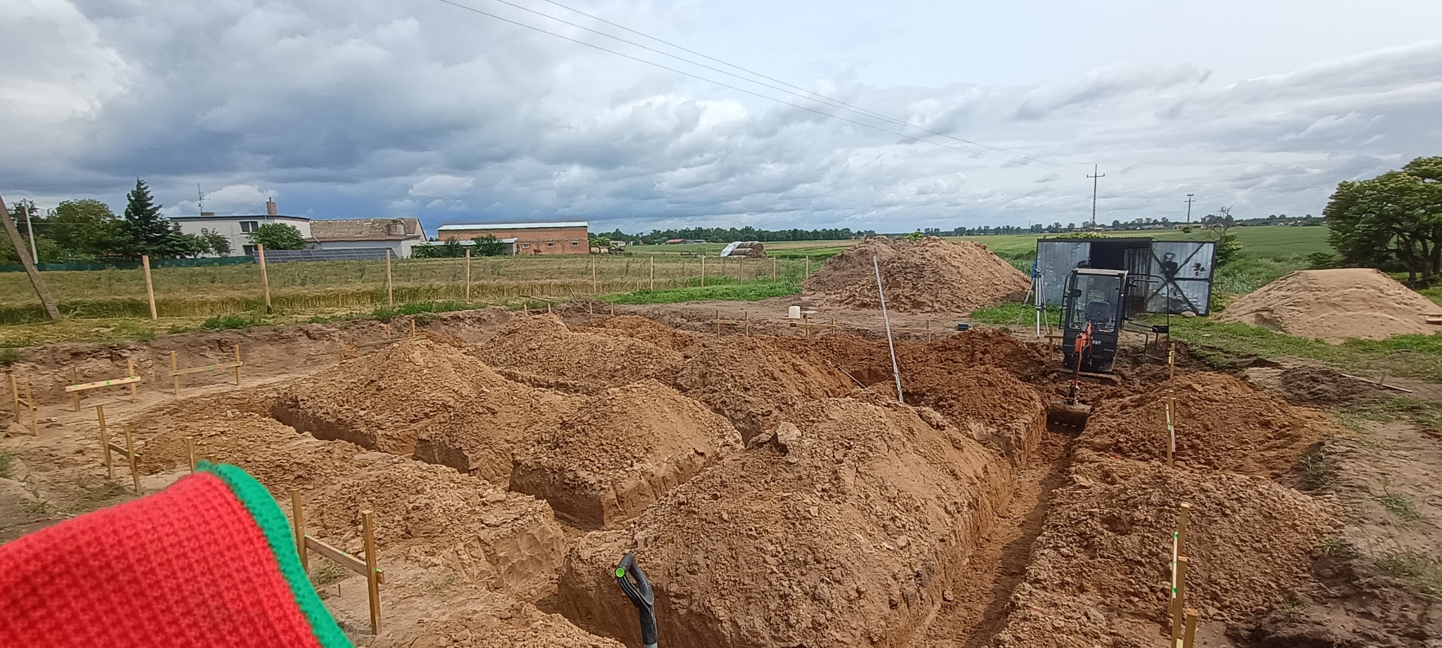 Wykopane rowy pod fundamenty z drewnianym szalunkiem, mała koparka i hałda ziemi w tle, pochmurne niebo nad polami uprawnymi.