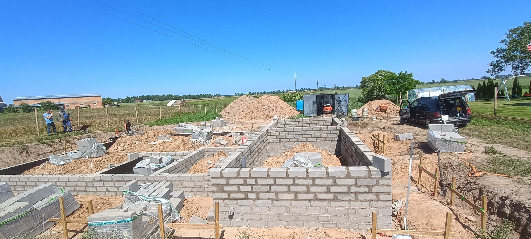 Budowa fundamentów z bloczków betonowych, widoczne narzędzia, betoniarka i auto. W tle zielone pola i błękitne niebo. Prace w toku, etap murowania ścian fundamentowych.