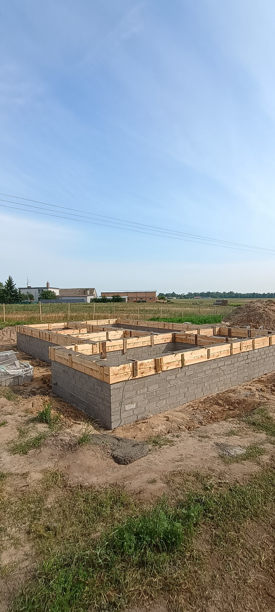 Fundamenty z bloczków betonowych, z drewnianym szalunkiem, na tle krajobrazu w Skokach. Widok z poziomu gruntu, błękitne niebo.