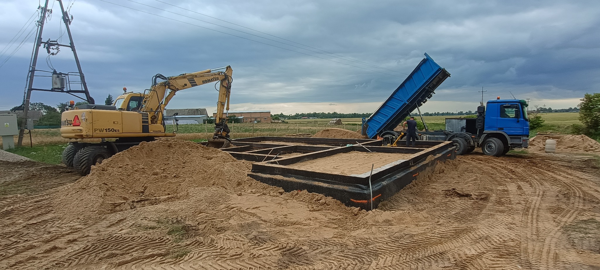 Żółty koparko-ładowarka Komatsu i niebieska wywrotka podczas zasypywania piaskiem fundamentów domu jednorodzinnego na piaszczystym terenie, pochmurne niebo.