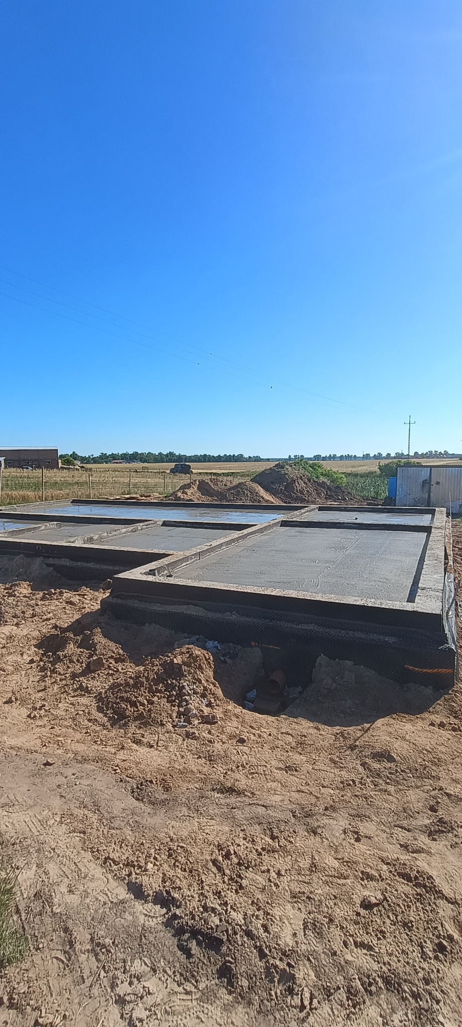 Zalane betonem fundamenty pod budowę domu jednorodzinnego, widoczne zbrojenia i izolacja. W tle pola uprawne i błękitne niebo.