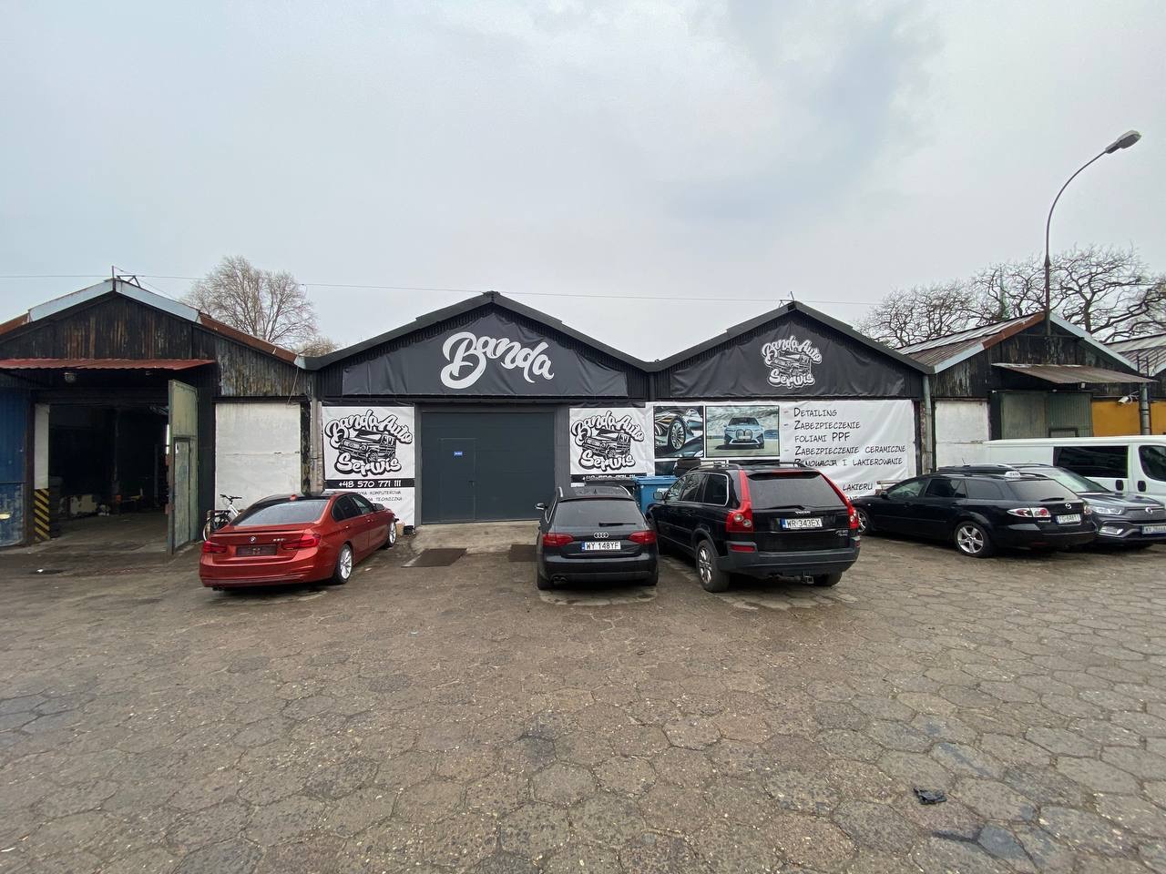 Szeroki kadr na fasadę warsztatu samochodowego 'Banda Auto Serwis' z zaparkowanymi samochodami. Na banerze wypisane usługi: detailing, zabezpieczenie foliami PPF, zabezpieczenie ceramiczne...