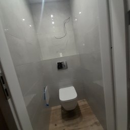 Perfect Marcin Roguski - Nowoczesna, wykończona łazienka z podwieszaną toaletą, szarymi płytkami ściennymi i drewnopodobną podłogą. Widoczne niedokończone podłączenia elektryczne.