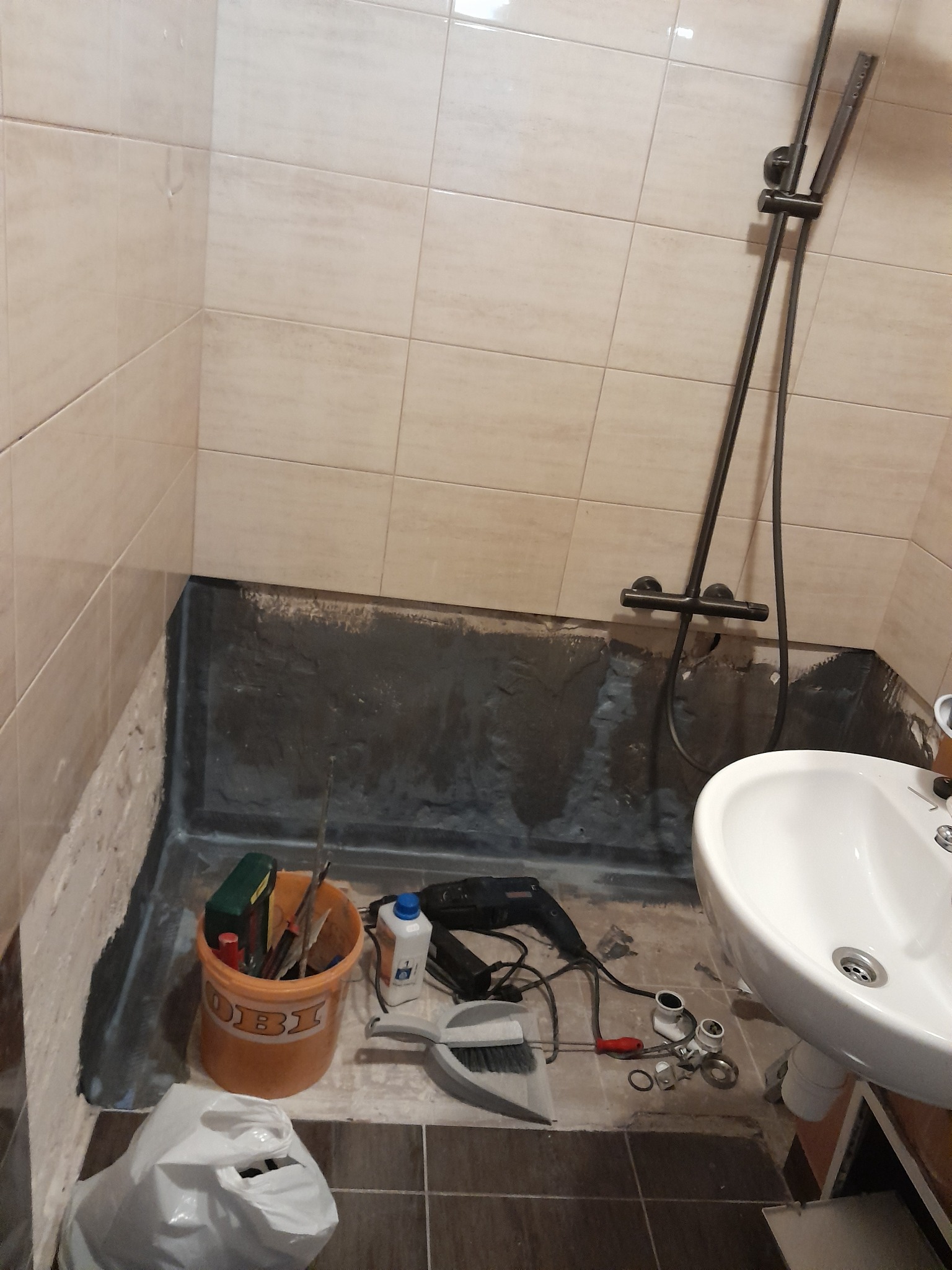 Przygotowanie do hydroizolacji w łazience: wiadro z narzędziami, wiertarka i akcesoria hydrauliczne obok umywalki, ściany z częściowo usuniętymi płytkami.