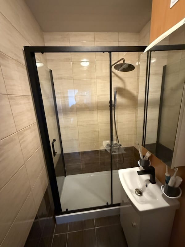 Nowoczesna kabina prysznicowa z czarną ramą i deszczownicą, umywalka z czarną armaturą, beżowo-brązowe płytki. Minimalistyczny design łazienki.